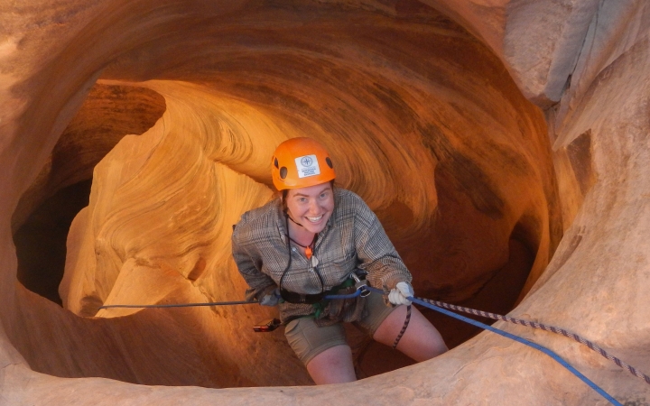 A person wearing safety gear smiles as they rappel into a canyon